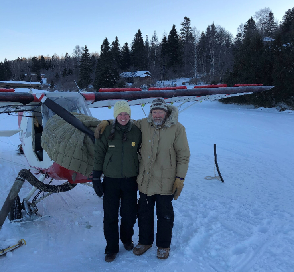 Out on the ice covered lake, a man and woman stand in front of a small plane. An island is nearby. a plane