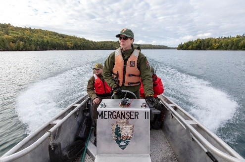 A woman in an NPS uniform wearing a PFD stands up while driving a boat called “Merganser II”.