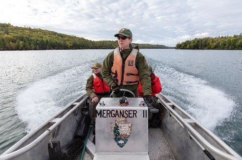 Driving a boat is part of a routine day’s work for staff at Isle Royale National Park. A woman in an NPS uniform wearing a PFD stands up while driving a boat called “Merganser II”.