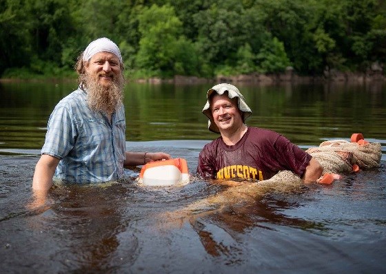Two men are standing waist or chest deep in a river.  One holds a rolled up net edged with orange floats.