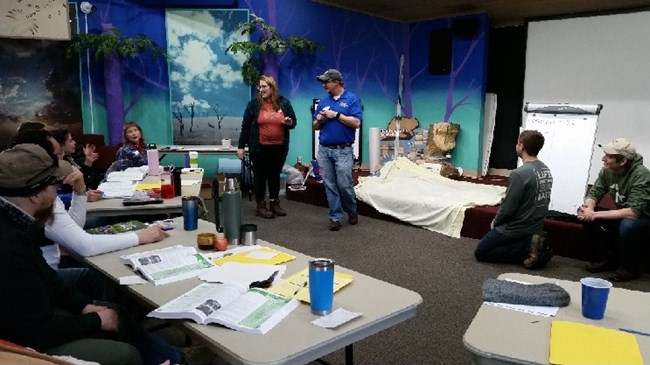 Small groups of educators tried their hands at leading the larger group in Project Wild activities during the 6-hour workshop. Several people stand or sit in the front of a room painted with a colorful forest scene while other people are seated at tables.