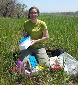 Researcher kneels at the edge of a wetland and prepares equipment for plant sampling.