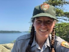 A woman wearing an NPS uniform smiles. A large lake and conifer are behind her.