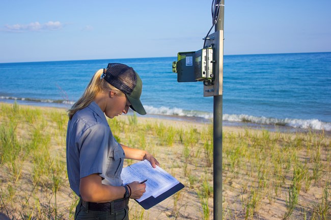 A woman in a National Park Service uniform stands next to a monitoring device mounted on a pole. She is recording data on a clipboard.