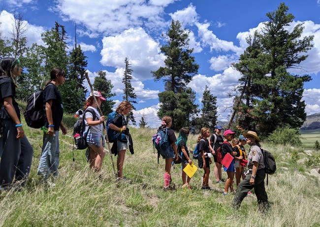 A park ranger addresses a large group of youth from a grassy hill overlooking a mountain landscape.