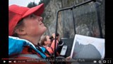 Still-frame of a video; a woman in a red hat looks up toward rocky cliffs.