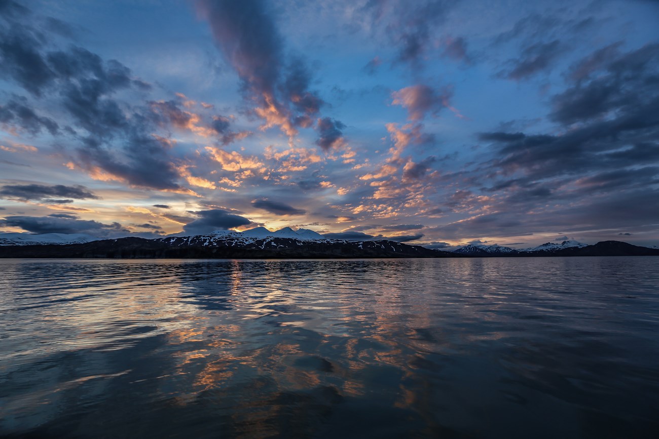 The sun sets behind coastal mountains; blue and orange clouds reflected in water in the foreground.