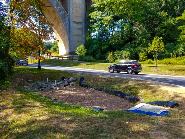 Low area with some large rocks next to resurfaced road and near large bridge.