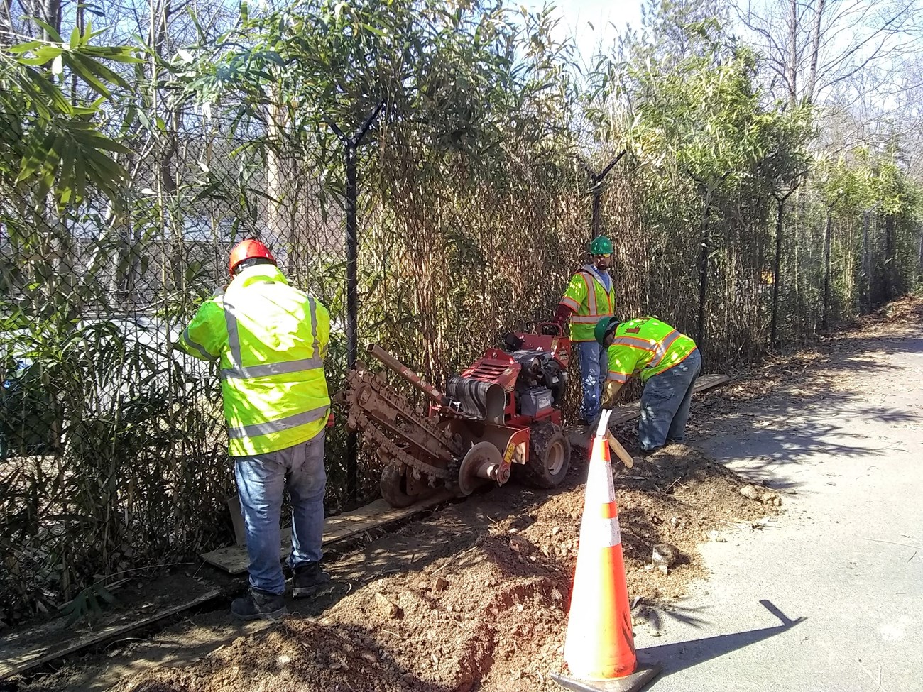 Three workers use machine to dig trench along a fence line between the Smithsonian National Zoo and Rock Creek Trail.