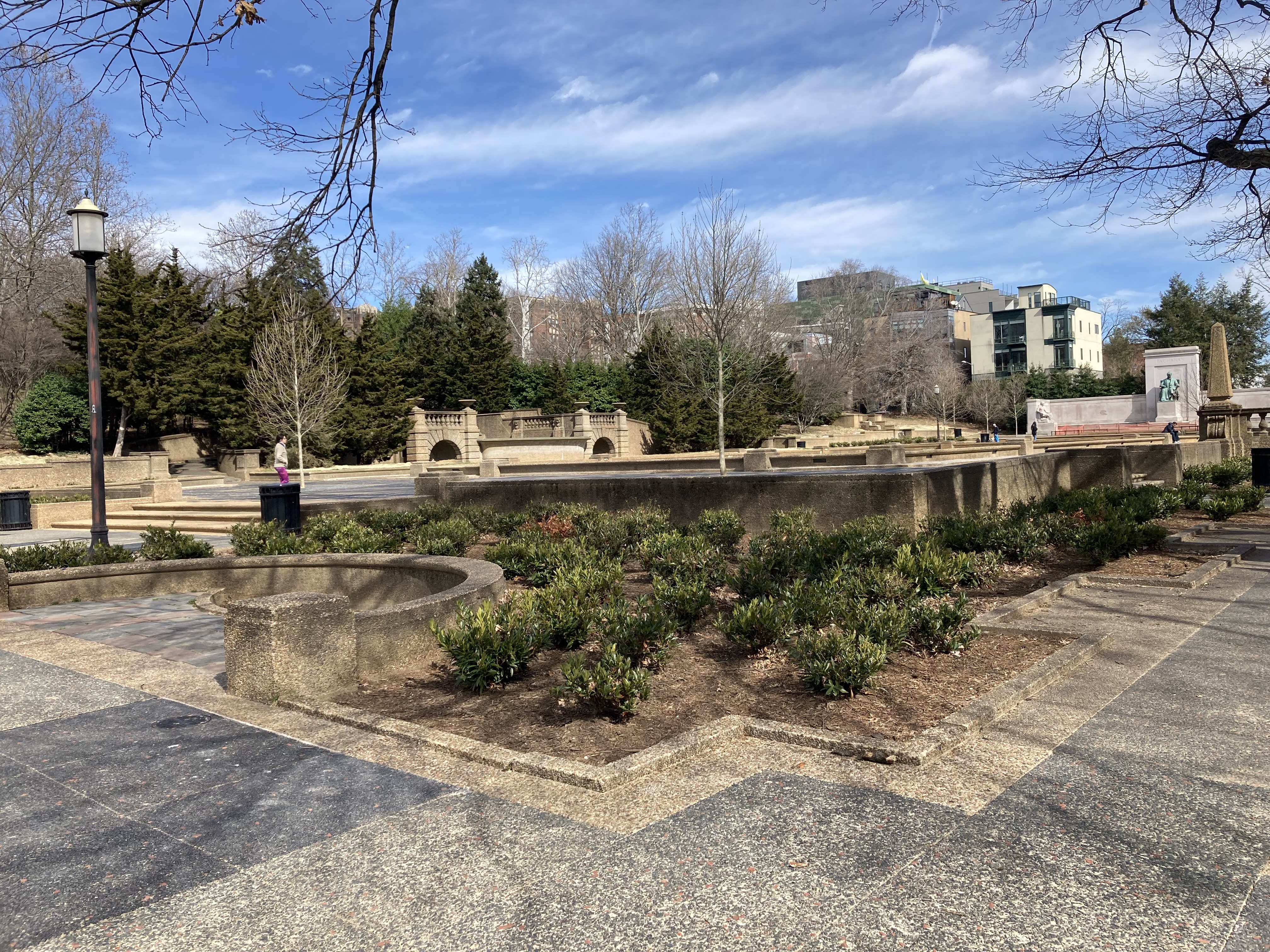 Stone pavers outline planters with shrubs in a broad plaza ringed with trees.