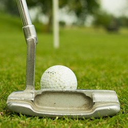 A close up of a putter putting a golfball on a green. The hole is blurry in the distance.