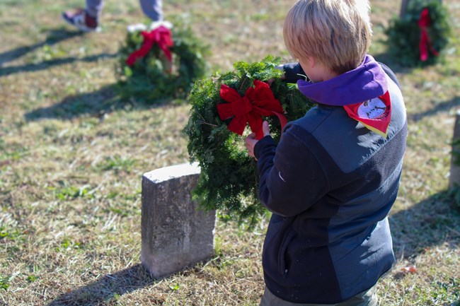 A young scout places a green wreath with red bow at a headstone