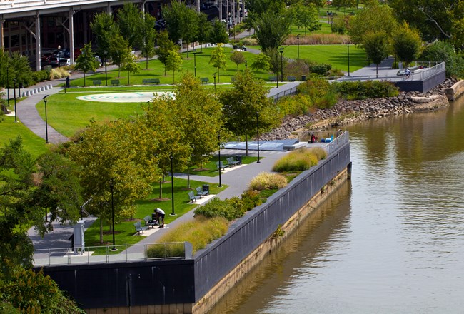 An aerial view of a sidewalk with a grassy area and trees to the left and a railing overlooking a river to the right.