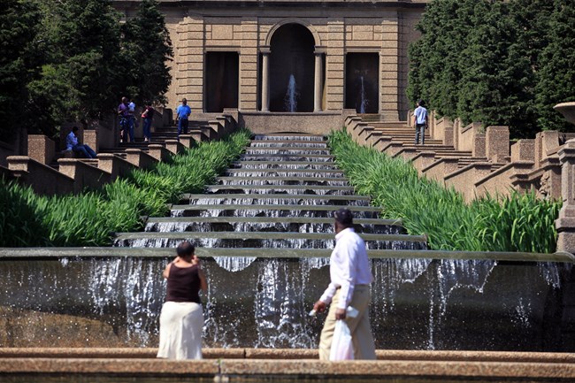A stone structure with a cascading fountain spilling downhill with grasses and stairs on both sides and two people looking at the fountain in the foreground.