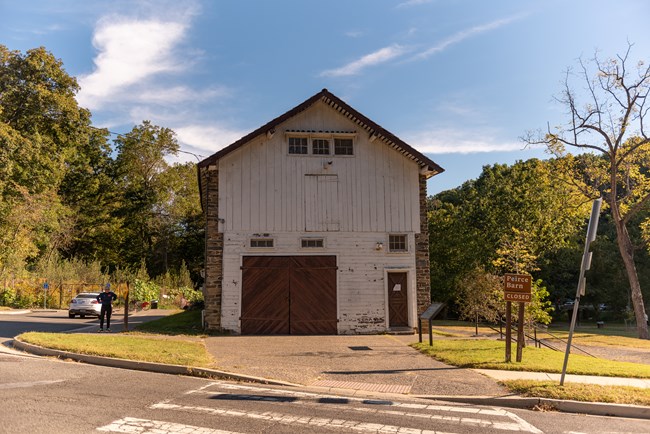 A street crosswalk leading towards a tall stone and white wood barn with trees behind it.