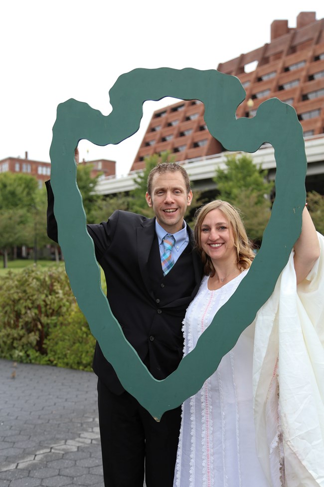 A man and woman in wedding attire hold up a wooden NPS arrow as a photograph is taken of them at Georgetown Waterfront Park in Rock Creek Park.