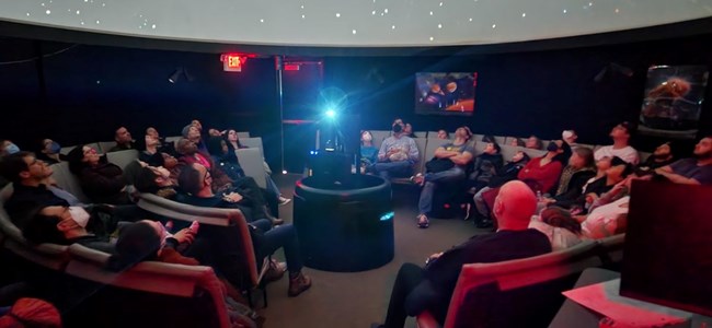 A group of people seated in a planetarium. They are looking up towards the ceiling at what the projector in the center of the image is projecting.