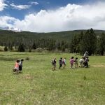 A line of students follow a park ranger through a green meadow with forests and tall mountains in the background and a blue sky with fluffy, white clouds overhead.