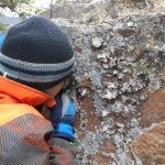 A student in a winter hat examines a rock closely with a magnifying glass.