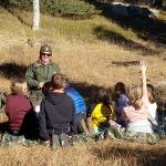 Students and a smiling park ranger sit in a meadow next to a ponderosa pine tree with a forest in the background.