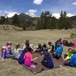 Students and a park ranger sit in a meadow with a forest and tall mountains in the background and a blue sky with fluffy clouds overhead.