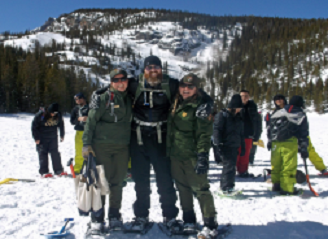 Two uniformed rangers stand side to side with teacher on a snowy hill with students in the background