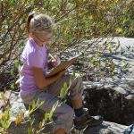 A student sits on a boulder next to bushes and writes in a journal.