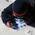 A student lays on snow and looks at snowflakes using a magnifying glass and a page with snowflake shapes.