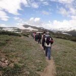 Students walk on a narrow trail through alpine tundra near snowcapped mountains.
