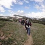 Students walk on a narrow trail through alpine tundra near snowcapped mountains.