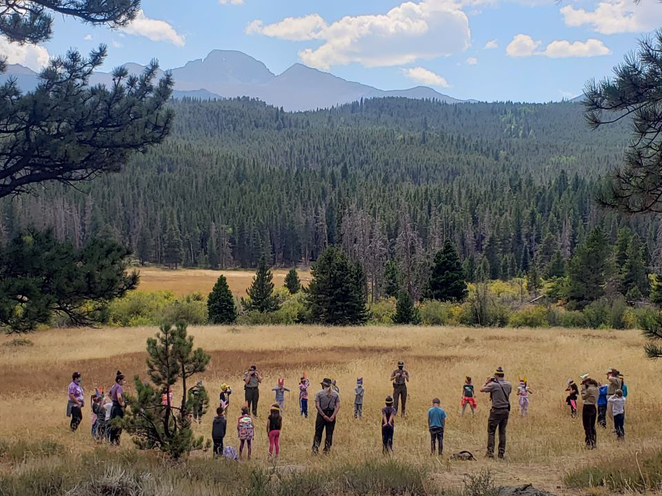 Students speak with a park ranger near a grassland and forest with tall mountains in the background.