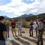 Students speak with a park ranger near a grassland and forest with tall mountains in the background.