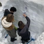 Three students stand in a recently dug snow pit and use tools to measure the layers within the snow.