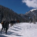Students snowshoe through a snow-covered mountainous landscape with a bright blue sky overhead and snow-capped mountains in the background.