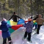 Students and an adult stand in a circle and hold the edges of an unfurled multicolored parachute while standing in a forest on snow.