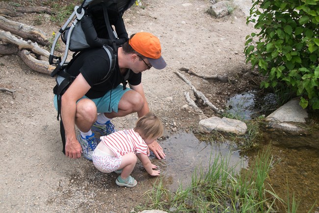 A father and daughter kneel down to explore the edge of a lake