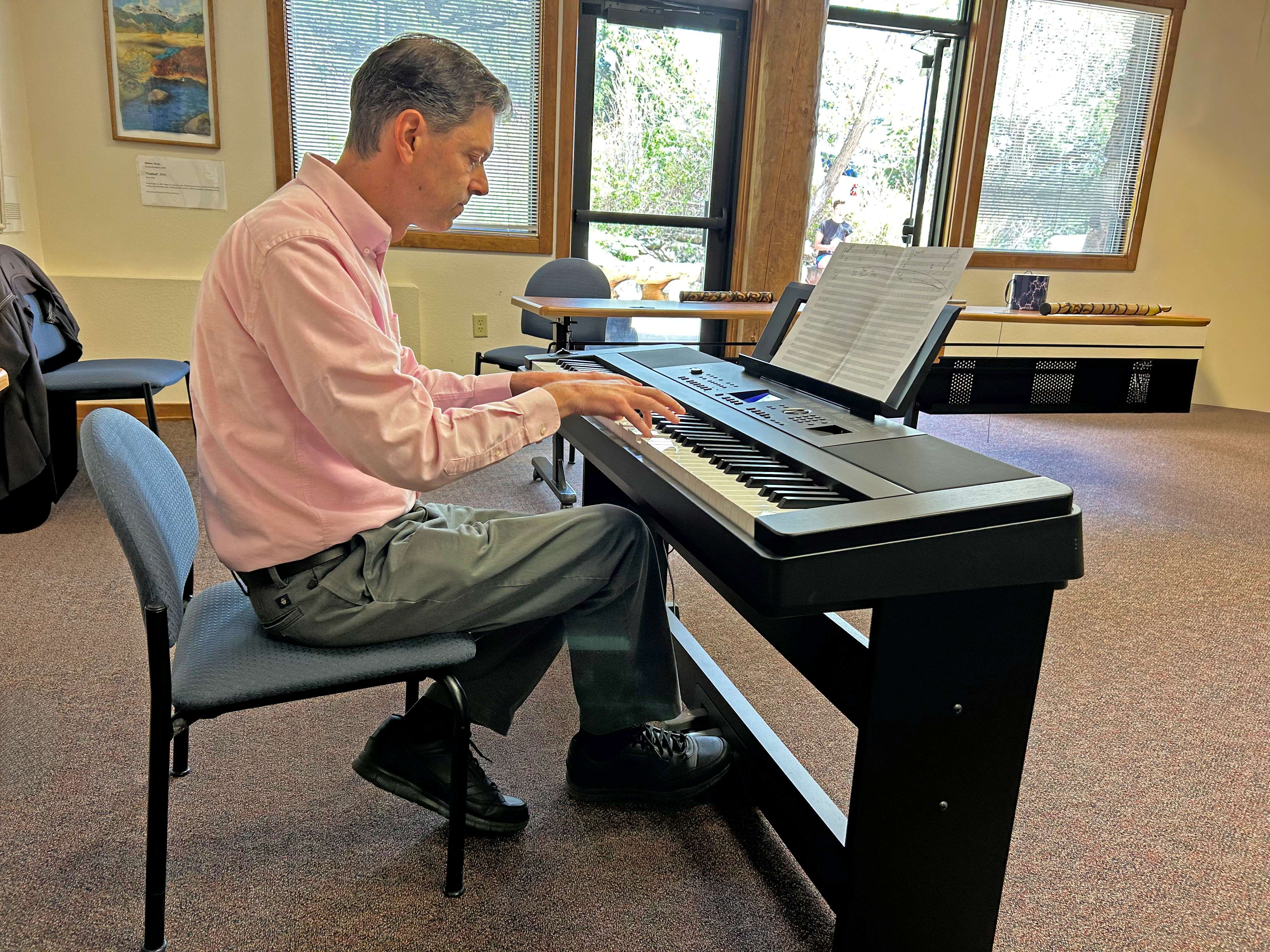 A man is sitting in front of an electric keyboard and he is reading sheet music. He plays a musical piece in a classroom.