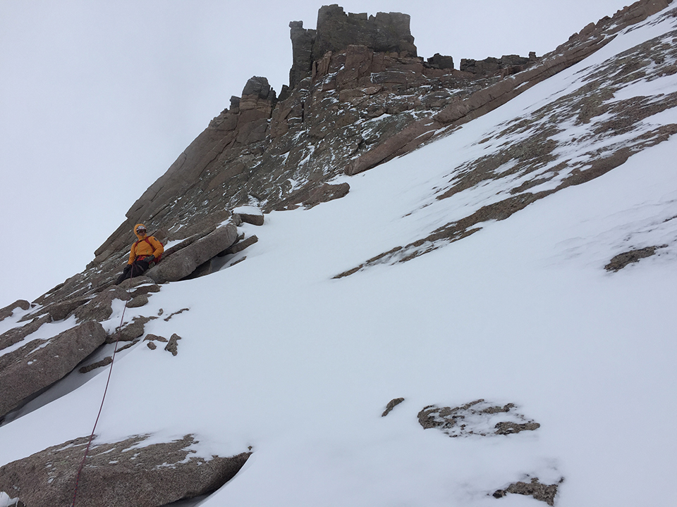 A climbing ranger, rope attached, sits on snow and ice on Longs Peak