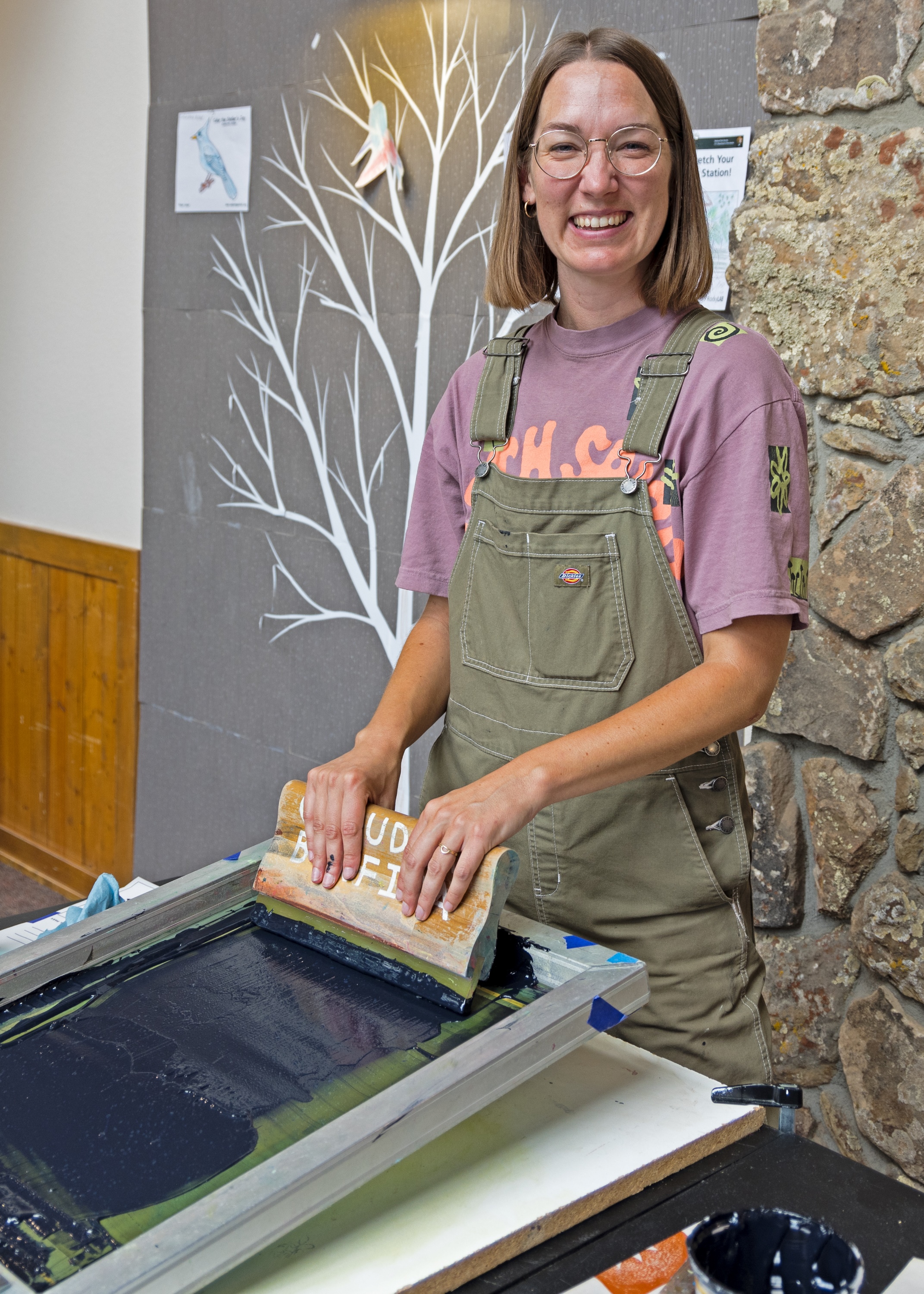 Visual artist Claudia Borfigua is smiling while wearing glasses, a purple t-shirt, and apron. A silk screen printing frame is on the table in front of her.