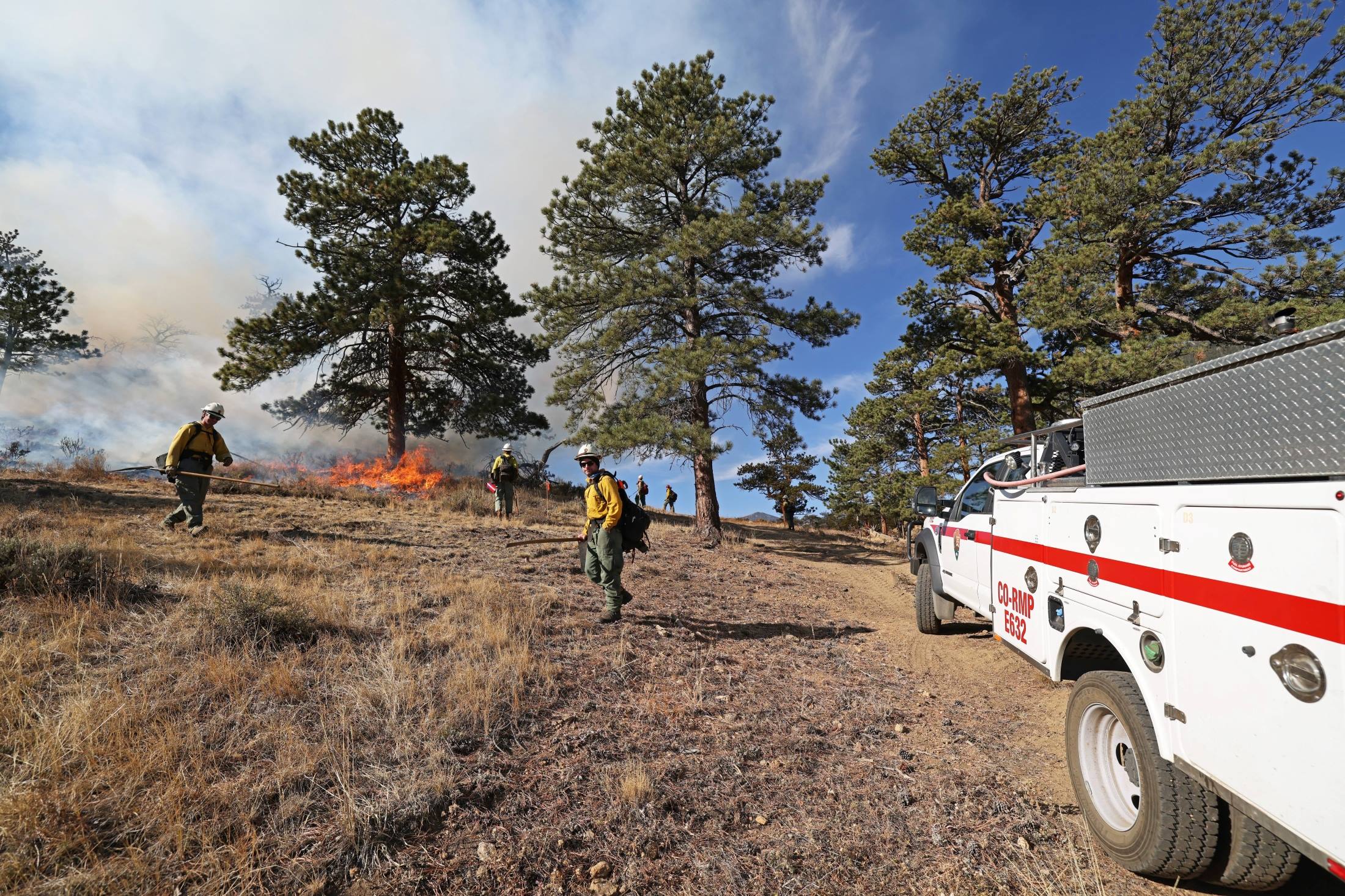 Courtesy RMNP_NPS firefighters and vehicle managing prescribed burn RMNP Firefighters are managing prescribed fire operations. A RMNP fire vehicle is present and a team of firefighters are carrying tools and managing fire on the ground as grasses and shrubs are burned.