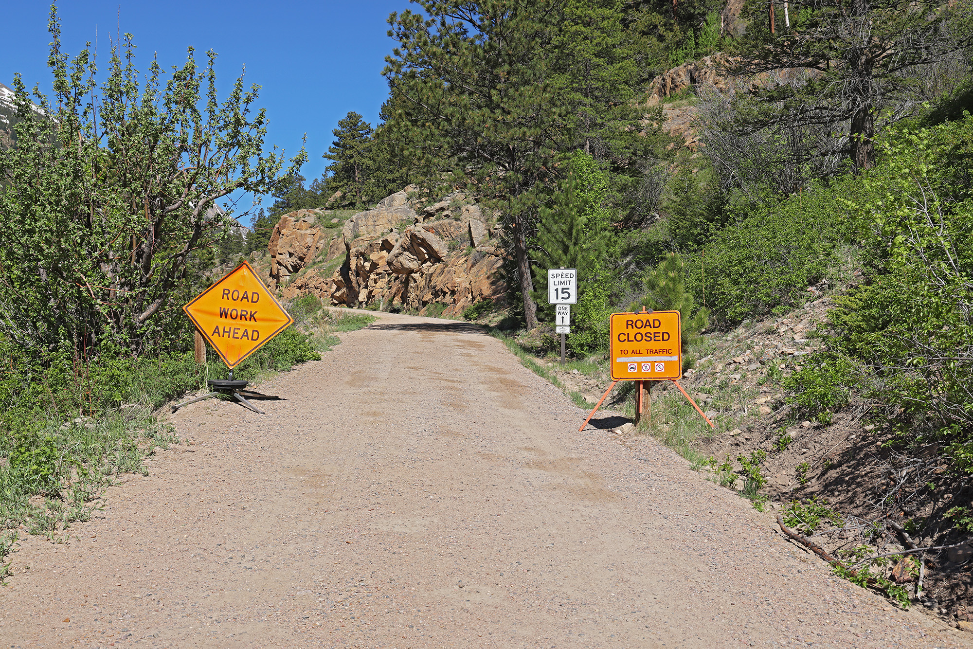 Courtesy RMNP_Old Fall River Road is Closed for Spring Opening Two orange signs are posted at the entrance to Old Fall River Road, a road with a dirt surface, showing the road is closed to all uses. Trees and vegetation line both sides of the narrow one-lane road on a sunny day.
