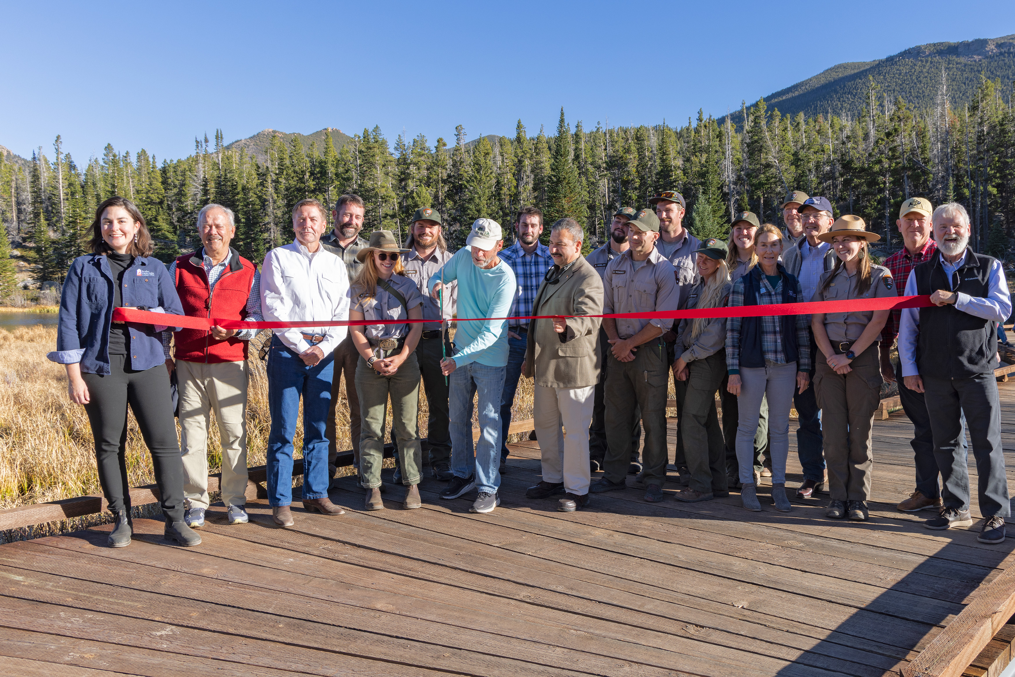 Ribbon cutting to celebrate the completion of the Sprague Lake Boardwalk Replacement Project. NPS staff and Rocky Mountain Conservancy Board Members and staff are gathered and holding a red ribbon across the boardwalk, to celebrate the completion of the Sprague Lake Boardwalk Project.