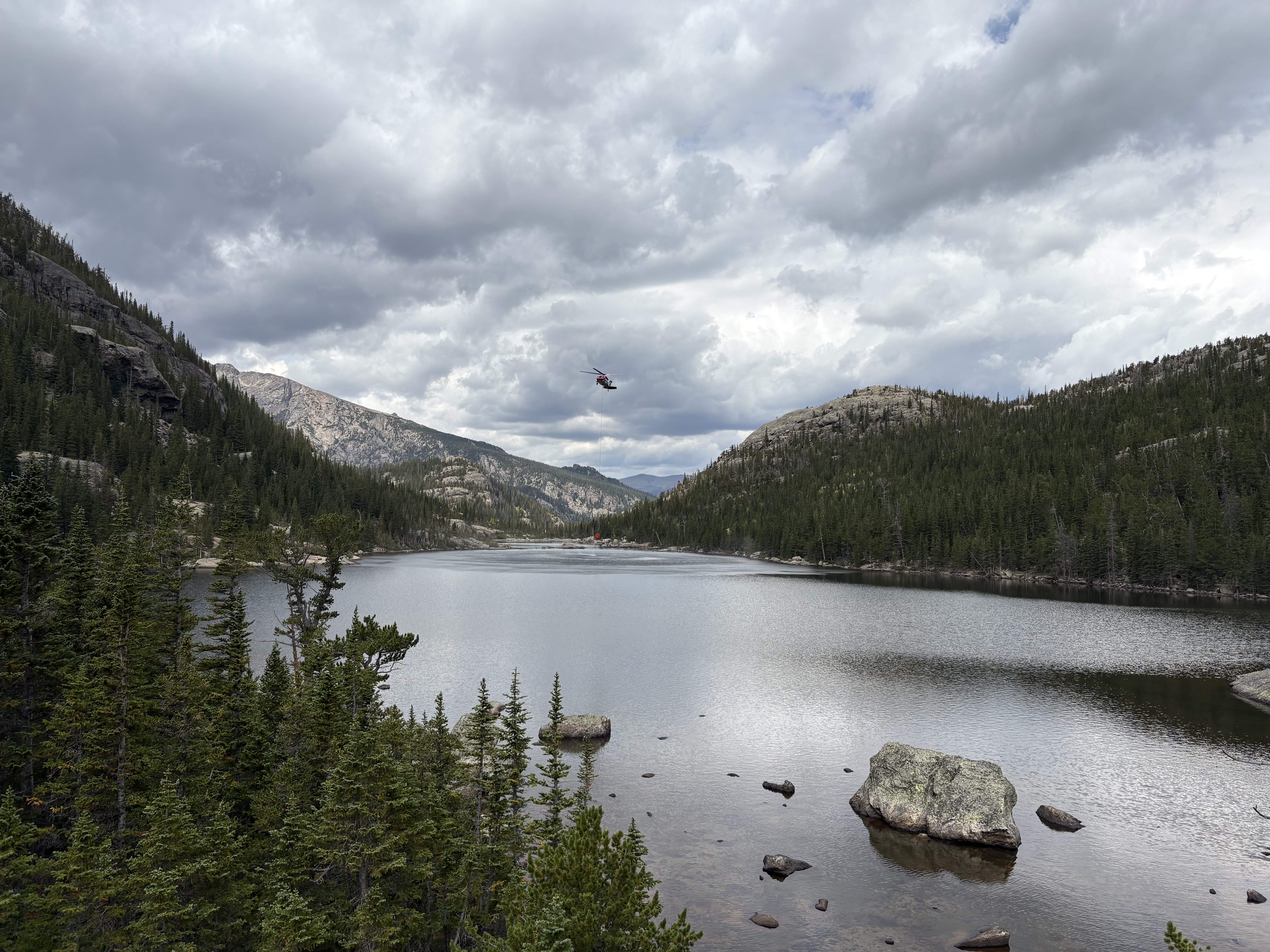 Helicopter with bucket for suppression efforts on the Jewel Fire (2) August 24 2025 A helicopter is dipping a bucket into Jewel Lake. The sky overhead is grey and cloudy.