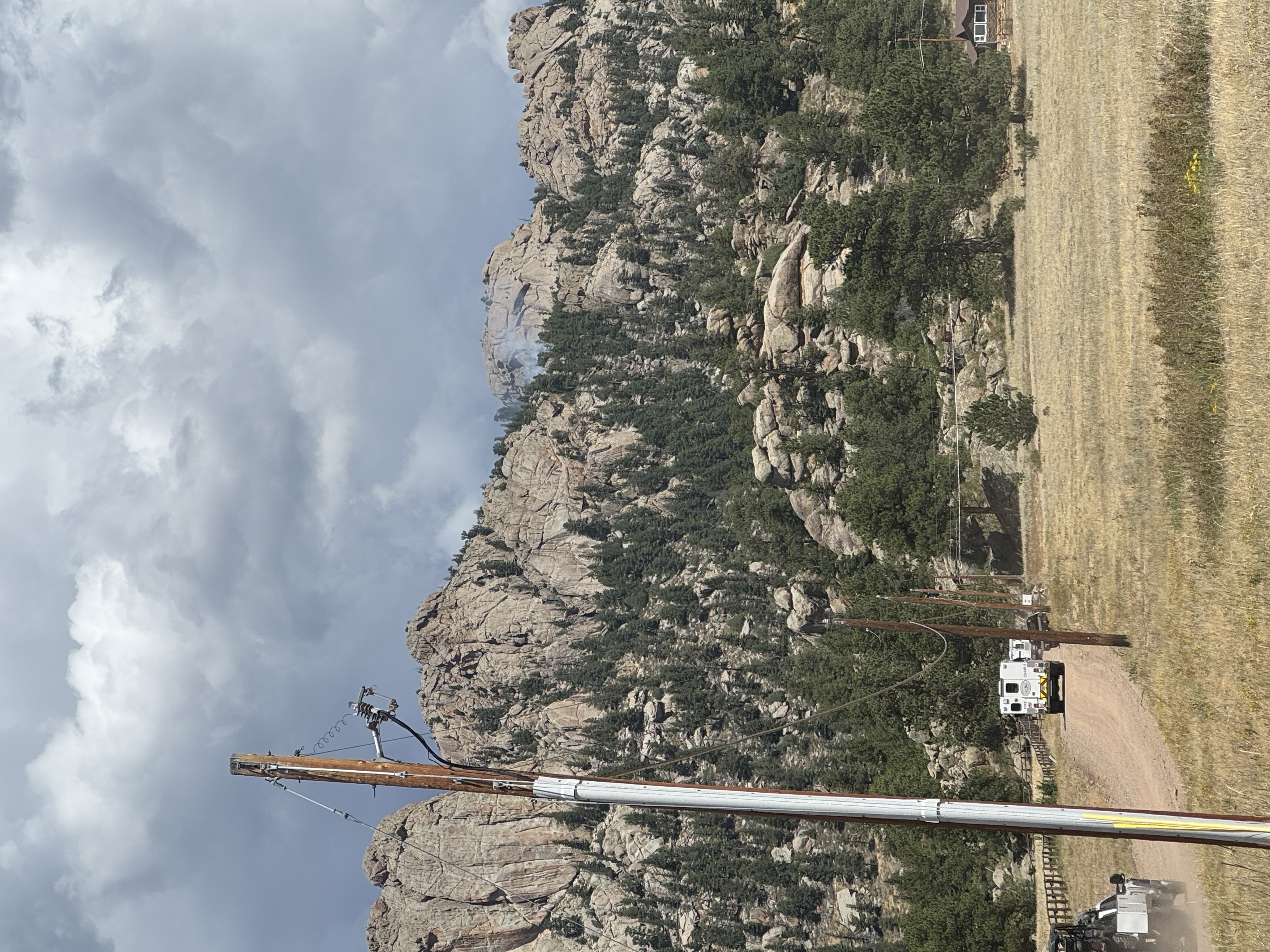 Smoke is rising from a forest hillside in RMNP. NPS fire trucks are enroute, driving down a dirt road outside the park. Power lines and homes are in the foreground. 
