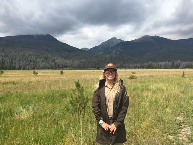 A woman wearing an intern uniform stands in front a grassy valley with mountains behind