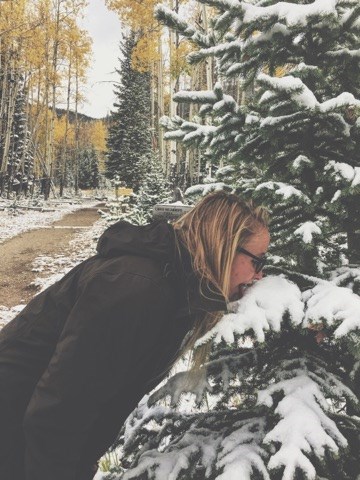 A woman stands with her face nearly touching a snow-laden tree branch with golden aspen in the background