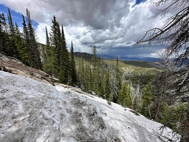 A steep section of the Lake Haiyaha Traverse is covered with snow on a cloudy day. There is a steep drop off to the right of the trail