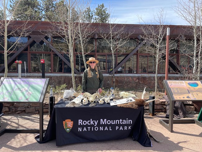 A park ranger in uniform is standing outside of the Beaver Meadows Visitor Center at a National Park Week activity booth