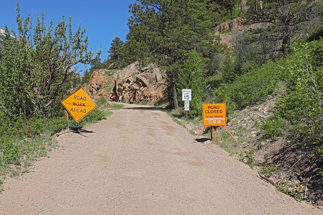 Two orange signs are posted at the entrance to Old Fall River Road, a road with a dirt surface, showing the road is closed to all uses. Trees and vegetation line both sides of the narrow one-lane road on a sunny day with clear blue sky