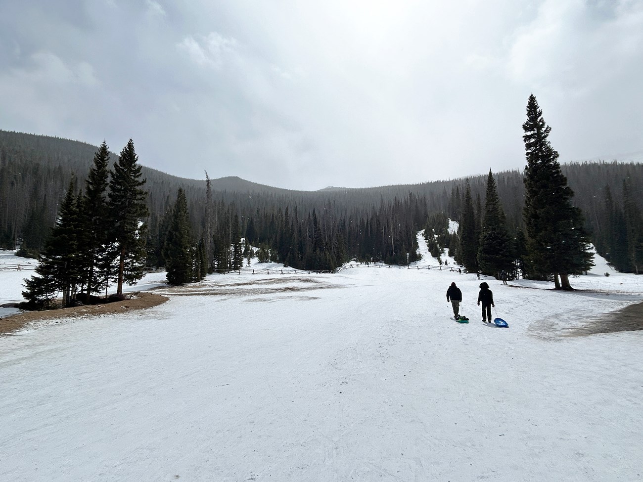 Right-side of the sledding hill is covered with a layer of ice and thin snow. Two people are walking sleds up the hill.